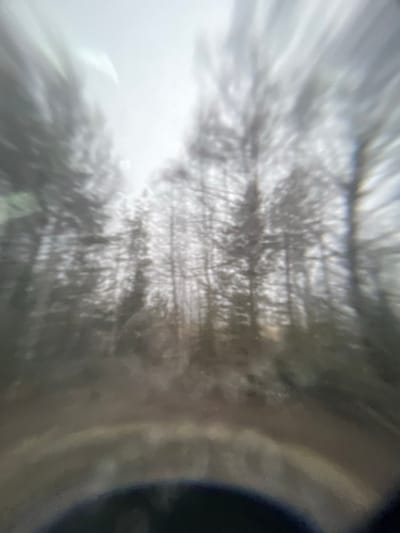 The image is blurry, showing a view through a lens, possibly a camera or optical instrument. The scene is dominated by grayish-white tones with some darker areas. The central area seems to contain multiple indistinct vertical lines, potentially representing trees or other similar objects. The right side features a curved dark area, suggesting part of the instrument's housing or structure. The overall image quality is poor, hindering precise identification of details. The location being Kukui, Russia is not discernible from the image content.