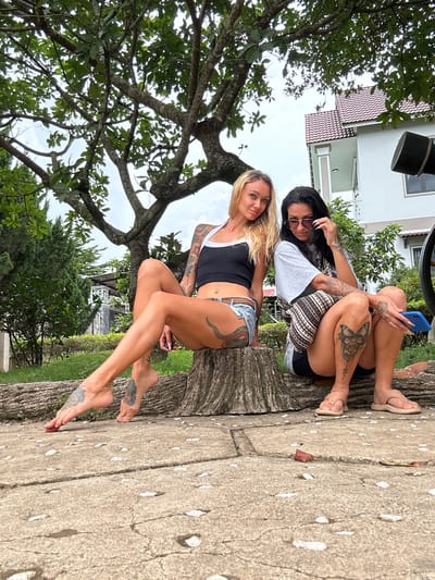 Two women are sitting on a tree trunk outdoors. The woman in the foreground, with blonde hair and tattoos, is wearing a black tank top and denim shorts. She is looking at the camera. The woman behind her, with dark hair, is wearing sunglasses, a grey t-shirt, and patterned shorts. She is holding a blue smartphone and appears to be looking at it. They are in a garden with trees and a building in the background. The ground is paved with concrete and has fallen flower petals on it. The image appears to be taken during the day, with overcast weather.