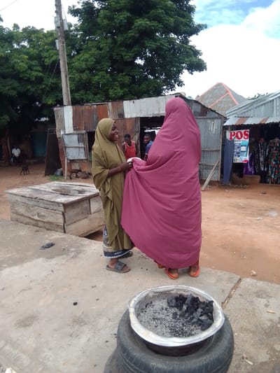Two women, both wearing headscarves, are engaged in a conversation outdoors in Gulum, Nigeria. The woman on the left, wearing a olive green headscarf, is smiling and looking at the other woman. The woman on the right is mostly obscured by a large, flowing magenta headscarf.
The setting appears to be a bustling market or street scene. Behind the women, there are several stalls made of corrugated metal. One stall has a sign that reads "POS". To the left of the women is a large, rustic wooden structure, possibly a well or a storage container. In the background, trees with lush green foliage can be seen, suggesting a warm climate. A power pole with electrical equipment stands tall behind the stalls.
In the foreground, a repurposed tire serves as a base for a metal container filled with charcoal and ash, indicating the presence of a cooking fire or brazier. There are also two small goats visible in the middle ground, adding to the rural atmosphere.
The lighting suggests it is daytime, with the sky appearing overcast. The overall mood seems pleasant, with the smiling woman conveying a sense of warmth and interaction. The scene depicts a slice of everyday life in a Nigerian community.