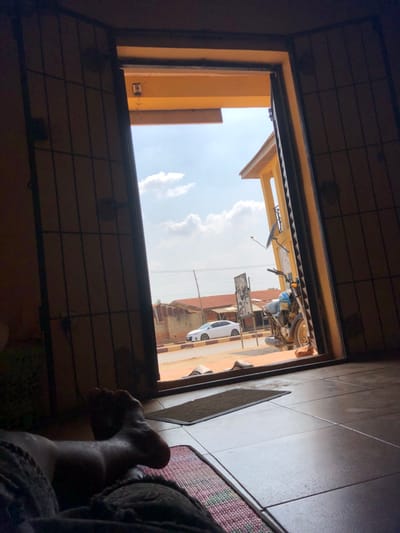 The image depicts an indoor perspective looking out through an open doorway onto a street scene in Ijara, Nigeria, during daylight hours under a partially cloudy sky.
In the immediate foreground, the feet and lower legs of an individual are visible on the lower left, resting on a patterned mat. The indoor floor consists of light-colored tiles, with a dark doormat situated directly inside the threshold. The doorway itself is composed of dark, grid-patterned metal security doors or grates set within a lighter, possibly yellowish-orange, frame.
Beyond the doorway, on a concrete step, two pairs of light-colored slippers are placed. To the right of the doorway, a blue or grey motorcycle with gold-colored wheels is parked on a kickstand. Behind the motorcycle, a dark rectangular sign or billboard stands near the roadside. Adjacent to this, a multi-story building, painted in a similar yellowish-orange hue to the door frame, is visible on the right, featuring a balcony and a satellite dish mounted on its exterior.
Further into the background, a paved road stretches horizontally across the scene. A white sedan is parked on the opposite side of the road. Beyond the road, several low-rise buildings with reddish-brown roofs are visible, along with utility poles and overhead lines. The sky is bright blue with scattered white cumulus clouds.