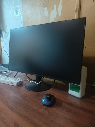 The image shows a desk with a computer monitor, a mouse, a power strip, and a white bottle. The monitor is black and has the AOC logo on the front. The mouse is blue and black and has the Logitech logo on it. The power strip is white and has multiple outlets. The bottle is white and has a green label with text in Russian. The desk is made of dark wood. The background is a wall with textured wallpaper and some exposed wood. The scene is indoors. This scene is not in Redkino, Russia.