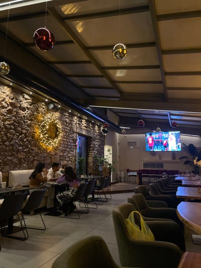 This media file captures an indoor scene, likely a restaurant, bar, or cafe, in Chaidari, Greece, during the evening or night. The image is oriented vertically (rotated 90 degrees counter-clockwise), but describes a space viewed from an elevated perspective looking down and across.
The setting features a distinctive design blending industrial and rustic elements. Overhead, a sloped ceiling made of light-colored panels and metal beams suggests a modern construction. Along one side, a prominent wall is constructed from irregular, dark-colored natural stones, providing a rustic contrast.
Decorative elements include several large, reflective spherical ornaments (some golden/silver, others deep red/burgundy) suspended at varying heights from the ceiling, casting subtle reflections. On the stone wall, a large, circular arrangement of glowing fairy lights creates a warm, wreath-like focal point. Other smaller red light fixtures are mounted along a structural beam. Greenery in planters is visible along the top of the stone wall.
Furniture in the establishment includes rows of modern chairs with black metal frames, some appearing to have dark upholstered seats, arranged along wooden-topped tables. A white, cushioned bench or sofa is also visible.
Several people are seated at tables, engaged in conversation or looking at their phones. In the foreground (bottom right of the original image), individuals are seated at a white bench. Further back, a person wearing a purple top and patterned skirt is visible.
A large flat-screen television mounted high on a wall displays what appears to be a football (soccer) match, showing players in red and blue jerseys. Faint text such as "ART" and numbers like "29" are discernable on the screen. The overall ambiance is warm and relaxed, illuminated by artificial lighting, suggesting a social gathering spot.