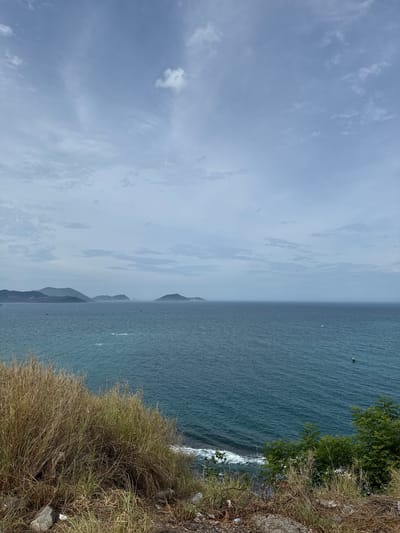 This is an eye-level, full shot of the coast in Nam Nha Trang, Vietnam. In the foreground, dry, yellowed grasses and a few rocks line the edge of a cliff overlooking the ocean. The sea is a deep blue, with white waves crashing against the shore. In the distance, several islands are visible, their slopes covered in vegetation. The sky is cloudy, with a few wispy clouds scattered across a pale blue expanse. A small, green boat is visible on the water in the distance.
