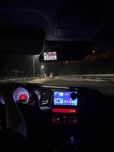 An interior view of a vehicle, likely a Kia Optima based on the dashboard design, is captured at night from the passenger's perspective, looking forward. A person's left arm and hand are visible on the steering wheel, indicating they are operating the vehicle. The speedometer shows a speed of approximately 80 km/h, the ambient temperature is 10°C, and the odometer reads 333081 km. The dashboard is illuminated with red and blue lights, including the instrument cluster, climate control buttons, and the hazard light switch. A large infotainment screen displays a car icon, various application icons, and the time 21:00. A separate red digital display below the infotainment screen shows 21:50. A dashcam is mounted near the rearview mirror, displaying a real-time feed of the road ahead, characterized by purple and green hues due to night vision or specific display settings. Through the windshield, a multi-lane road is visible, illuminated by streetlights. Guardrails line the right side of the road. The taillights of several distant vehicles are discernible. A tunnel or overpass structure appears further down the road. The scene is located in or around Chvizhepse, Russia.