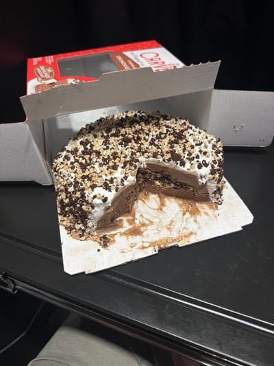 A chocolate ice cream cake with white frosting and chocolate and peanut toppings is sitting in an open cardboard box. The cake has a slice removed, and there is some frosting and cake debris around it. The box is on a dark, reflective surface, possibly a table. The background is dark and out of focus.