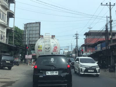 This media file shows a street scene in Palohlada, Indonesia, during the daytime under an overcast sky. The scene is dominated by a traffic jam, with several vehicles visible, including a black SUV with license plate BL 1168 NR, a white MPV with license plate B 2243 SRE, and a large white tanker truck. The tanker truck has text that reads "15.000 KG PT. MARWAH GAS BADI" and "PERTAMINA CONTACT CENTER". There are also signs for "AMPERA MINANG" and "NASI SATANG PADANG", indicating food establishments.
The image captures a typical urban environment with overhead electrical wires and utility poles. The buildings on the sides of the street appear to be a mix of commercial and residential structures. The traffic light in the distance is red, contributing to the congestion. One person is visible riding a motorcycle in the background. The overall mood of the image is one of everyday life and activity in a busy Indonesian city. The weather appears to be cloudy, suggesting it might be humid or raining.