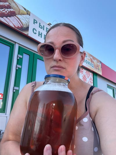 A woman with fair skin and light brown hair pulled back is holding a large glass jar filled with a brown liquid, possibly homemade beer or kombucha. She wears round pink sunglasses and a light pink polka dot top. The background shows a storefront with Cyrillic text, likely in Grigorovo, Russia. The text includes "РЫБ" and "МОНАА," suggesting the store sells fish and possibly beer. The storefront has green trim and windows. The sky is light blue.