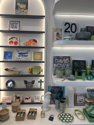 The image displays an interior retail scene from a homewares and gift boutique located in Bondi Beach, Australia. The environment is well-lit with integrated strip lighting illuminating multiple shelves against a white wall.

On the left, an arched shelving unit with dark shelves holds various decorative items. From top to bottom: a framed "MOJITO" artwork; a framed artwork with "L'HEURE DE LE SIESTE" text; a square artwork featuring two cherries and a separate, orange-toned seashell-shaped artwork; three square artworks designed like vintage postage stamps (depicting a seashell, two sardines with "SARDINIA" text, and a sun with "16 LIRE" text); two fish-shaped artworks (one yellow and blue, the other yellow, blue, and green); a small circular mirror, a butterfly-shaped artwork, and a blue cross-shaped artwork; and four small square artworks depicting various dogs (a Dalmatian, a tan dog, a Basset Hound, and a white horse).

On the right, a series of white, curved shelves extends. From top to bottom: a book titled "20," a framed "50 LIRE" Campari postage stamp artwork, a green vase, a white bracelet with "WORK OF ART" text, and other books. The shelf below holds a "DAVID BOWIE" book, a "PLANTO" book, two green and white striped vases, a green glass teacup and saucer, a set of green candle holders, a green pear-shaped ornament, and a green pumpkin-shaped ornament.

The bottom area features a white display counter. On the left are two light brown, ruffled ceramic pots with matching saucers and two small brown boxes of "Assemble" brand chocolates. In the center are a white ceramic rooster-shaped pitcher, a small gold-colored jar, a blue tag attached to a light-colored ring, three tall green and white striped cylindrical vases, and a box of white taper candles. To the right are a green polka-dot plate with a wavy edge, an oval plate with a palm tree design, a stack of green and white striped plates, a brown box labeled "GRAND DESIGNS," and a mushroom-shaped decorative object. No people are visible.