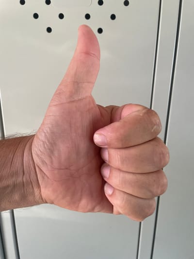 A close-up, side-profile shot shows a person's right hand with the thumb extended upwards in a "thumbs-up" gesture. The hand is positioned in front of a light-colored, textured surface that appears to be part of a locker bank, with vertical lines indicating seams or doors. The background is out of focus, but the matte white surface with black dots is visible. The image is well-lit, with natural light casting soft shadows on the hand and highlighting the wrinkles and skin texture. The arm and wrist are partially visible, with short, dark hairs on the forearm.