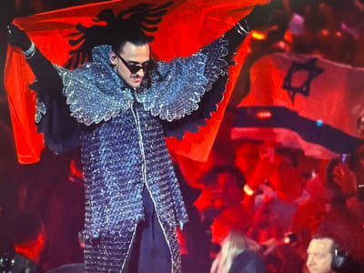 The image depicts a person in a highly decorative, sparkly outfit, holding two flags. The person is wearing dark sunglasses and has black hair. The outfit appears to be a jacket with wide, shimmering lapels and possibly a cape or train that extends downwards. The person is positioned upside down in the frame, with their head at the bottom.
One of the flags is predominantly red with a dark six-pointed star (Star of David) in the center and a horizontal blue stripe above it. This is the flag of Israel. The other flag is solid red, with a black double-headed eagle insignia. This is the flag of Albania.
The background is a blur of red and some lighter shapes, suggesting a crowd or audience. The overall impression is that of a performance or event, possibly a concert or a public gathering, where flags are being displayed. The lighting is dramatic, with bright highlights and deep shadows, creating a theatrical atmosphere. The context provided is Ellwangen, Germany, which could indicate the location of this event. The emotion conveyed is difficult to determine due to the sunglasses and the upside-down orientation, but the elaborate costume and the display of national flags suggest a celebratory or significant moment. There is no visible text in the image.