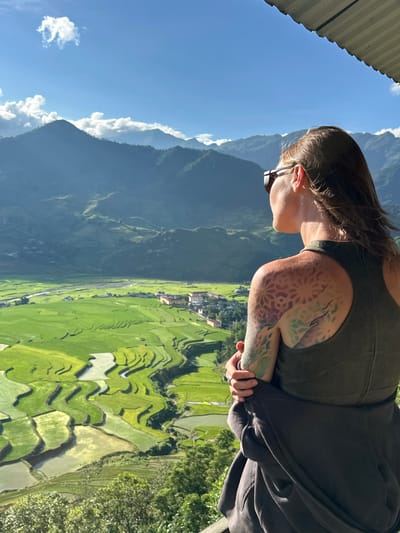 A person is observed from the back, facing away from the camera, looking out over a verdant valley in Tú Lệ, Vietnam. The individual, a female, wears dark sunglasses, a dark olive green tank top, and has a dark gray jacket tied around her waist. A detailed, colorful tattoo featuring geometric and organic patterns is visible on her left shoulder and upper arm. Her brown hair is parted on the left and falls over her shoulders.

The expansive landscape before her consists of rolling mountains under a bright blue sky with scattered white clouds. In the foreground and midground of the valley, extensive terraced rice fields display varying shades of bright green and pale, water-filled sections, indicating different stages of cultivation. A river winds through the valley, passing near a cluster of buildings that form a small village or town. The sunlight casts clear shadows, highlighting the contours of the mountains and the intricate patterns of the rice paddies. The top right corner of the image shows a portion of a corrugated metal roof or overhang.