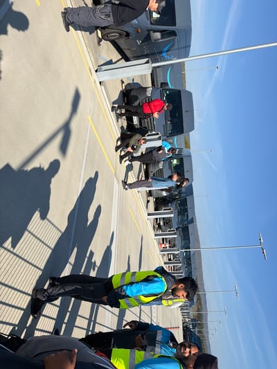 The image shows a parking lot in Friedrichsdorf, Germany, filled with gray Amazon delivery vans. Several people are standing or sitting near the vans. Some are wearing vests and turbans and some are wearing red coats, seemingly in conversation or taking a break. The scene is illuminated by bright sunlight. Parking spaces are delineated by yellow lines. The lighting is clear and there are shadows of a fence in the foreground.