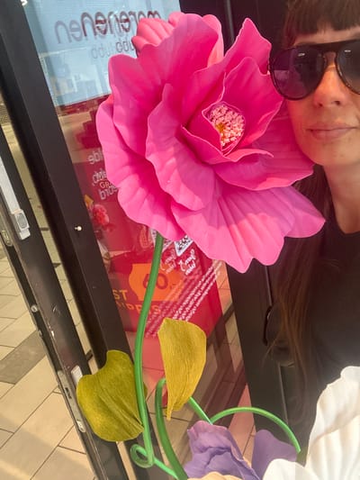 This media file shows a close-up, top-down view of a person lying down, with their face partially obscured by sunglasses and a large, bright pink artificial flower. The person has dark hair and appears to be resting or sleeping. The setting is outdoors, as suggested by the sunlight reflecting off surfaces and the warm lighting.

The most prominent object is the oversized pink flower, which has delicate, layered petals and a textured center. Beside it, part of a floral arrangement is visible, including light purple and white artificial flowers and green stems. These flowers are positioned on a reflective surface, likely a table or window, which displays subtle reflections of buildings and possibly text.

Beneath the flowers, a red book or magazine is visible, with some text printed on it, though it's largely illegible due to glare and the angle. One section of text in white lettering on a light background on the left side of the image appears to be a logo or brand name, possibly "nenenexu." Other visible text elements are scattered and unclear.

The overall impression is one of a peaceful moment, possibly a nap taken outdoors on a sunny day, surrounded by decorative artificial flowers. The image has a bright and cheerful color palette dominated by the vibrant pink of the flower. The time of day appears to be late morning or afternoon, given the quality of the light. The location context of Slitu, Norway, does not provide specific visual cues within the image itself.
