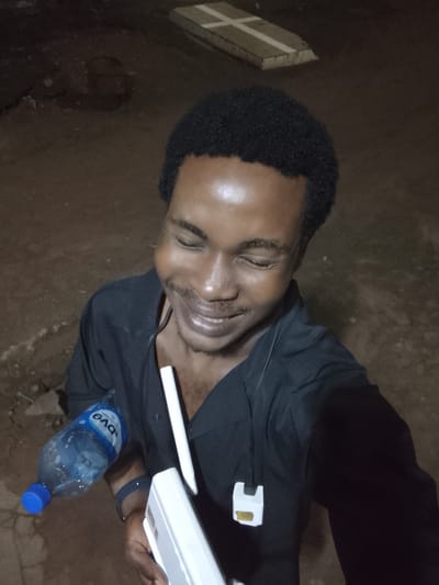 This is a medium-shot, slightly high-angle, self-portrait of a young Black man taken at night in Orodo, Nigeria. The man is smiling with his eyes closed, and his face is illuminated by what appears to be artificial light, creating a highlight on his forehead and cheek. He is wearing a black collared shirt. In his left hand, he holds a clear plastic bottle of water with a blue cap and a white label that reads "EVO" in blue letters. In his right hand, he is holding a white object, possibly a remote or a small device, and a white stylus or pen. A white cord hangs from his neck and is tucked into his shirt. In the background, the ground is dirt and appears uneven, with some debris. In the upper right of the frame, a portion of a concrete structure with two white stripes, resembling a cross or marker, is visible. The overall setting appears to be outdoors at night, possibly in a less-developed area given the terrain and the presence of the concrete marker, which could be a grave or part of a building foundation. The man's expression conveys happiness and relaxation.