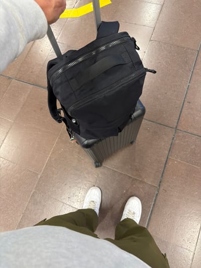In Stockholm-Arlanda Airport, a person stands with their luggage. A black backpack is placed on top of a dark-colored rolling suitcase. The person is wearing white sneakers and olive-green pants, with a light-colored hoodie visible. The flooring is tiled with brown and tan squares. A yellow line is visible in the background.