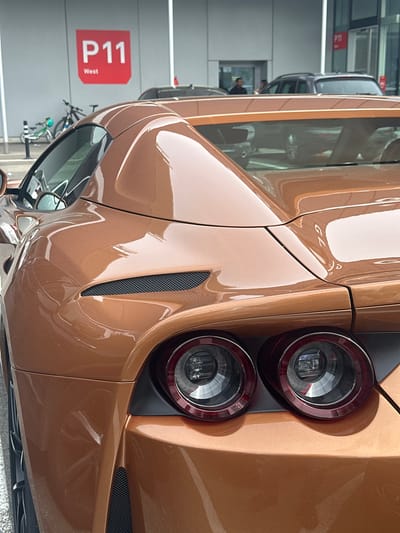 A striking bronze-colored sports car is the central subject, captured from a low rear-three-quarter angle in what appears to be an indoor parking facility or showroom in Wallisellen, Switzerland. The car's highly reflective metallic paint gleams under artificial lighting, showcasing its sculpted lines. It features distinctive, modern circular LED taillights set within a dark housing, and a sleek black mesh vent is integrated into the side panel near the rear wheel arch. A faint reflection or emblem resembling "GL" is visible on the rear quarter panel.
The setting consists of concrete walls and structures typical of a multi-level parking garage. Reflections in the car's windows and glossy paintwork reveal parts of the environment: several other parked vehicles, including grey sedans or SUVs, a person in a dark shirt standing in the distance, and a rack with multiple bicycles. A prominent red sign in the background clearly displays the text "P11 West," likely indicating a parking level and section. The car is stationary, suggesting it is parked or on display. There are no direct indications of weather or time of day beyond the indoor lighting.