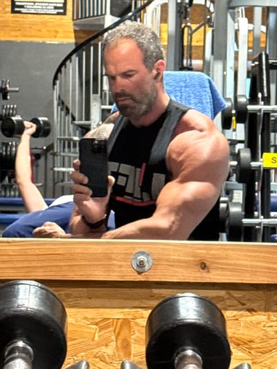 The image shows a man in a gym, presumably in Jemelle, Belgium, taking a selfie in a mirror. He is wearing a black tank top, holding a smartphone, and has gray hair and a beard. In the reflection, another person is visible lifting a dumbbell. The gym equipment and weights are scattered in the background and along the right edge of the frame. The mirror is mounted on a wooden surface.