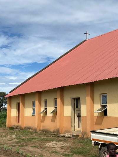 A low-angle shot captures a church building in Bawan Dodo, Nigeria. The church features a red corrugated metal roof, a tall, narrow white door with a brown cross, and several open windows. The walls are painted in alternating bands of yellow and orange. A small child in a pink shirt is partially visible in the lower right corner, standing next to the back of a white pickup truck. The ground around the church is a mixture of dirt and grass, with some scattered rocks and debris. The sky above is partly cloudy with a light blue hue.