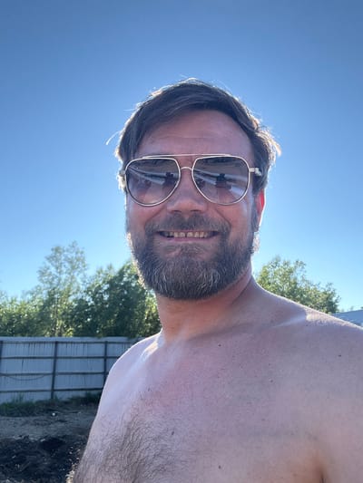 This media file shows a close-up, selfie-style shot of a man outdoors on a sunny day. The man is wearing aviator-style sunglasses and has a beard. His mouth is open in a smile, revealing his teeth. The sun is bright, casting a glow on his skin and hair, and causing some lens flare. The background consists of a clear blue sky, green trees, and a glimpse of a fence. The overall impression is one of a casual, happy moment captured in natural light, possibly during warm weather given the man's exposed shoulder and the bright sunshine. The image is oriented vertically, with the top of the man's head towards the right side of the frame. There is no visible text in the image.