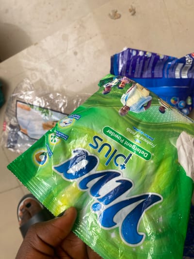 This is a close-up shot of a green and white packet of "Viva plus" detergent powder. The packet is slightly crinkled and appears to be held by a person's hand, with a foot wearing a sandal visible at the bottom right corner. The packet has large blue lettering spelling out "Viva plus". There are images of people on the packet, including what appears to be a family. Text on the packet indicates it is a "Detergent Powder" and mentions "freshness of petals" in both English and French ("fraîcheur des pétales"). There are also smaller icons and text highlighting features like "Colour Protection", "Multi-Stain Removal Tech", and "Removes Tough S". In the background, another blue detergent packet is partially visible. The setting appears to be indoors with a tiled floor. The lighting is bright, suggesting it is daytime. The overall impression is that of a product being displayed or handled.