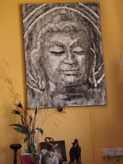 A large, textured painting of Buddha's face dominates the upper center of this indoor scene, hanging against a warm yellow/ochre wall. The painting is rendered in shades of black, white, and grey, featuring a textured, possibly iridescent finish that catches the light. Buddha's eyes are closed, conveying a sense of serenity, and concentric, halo-like rings are visible above the head.
Below the painting, on what appears to be a surface or shelf, is a collection of decorative items. A vase, partially visible, holds a mix of dried, curly branches, several peacock feathers, and a green leafy plant. To the right of the vase, various small sculptures are arranged. These include a dark wooden elephant figurine with its trunk raised, another wooden sculpture, and two small grey animal figurines, one resembling an elephant and the other possibly a sheep or ram.
The wall itself is a solid, warm yellow or ochre color, and appears to have a slightly textured finish. In the lower right corner, a white power strip with multiple sockets and a small red indicator light is mounted on the wall, with a white cable extending downwards from it. The lighting suggests an indoor setting, possibly during the day or evening with artificial illumination. The overall impression is one of a thoughtfully decorated space, potentially in a home or commercial establishment in Saint Paul's Bay, Malta. No discernible text is visible in the image.