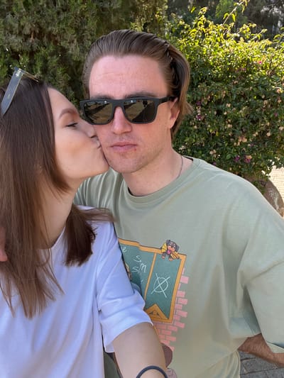 A couple is posing for a selfie outdoors. The man, wearing black sunglasses and a light green t-shirt with a dog graphic, is being kissed on the cheek by the woman. The woman has long brown hair, and is wearing a white t-shirt and sunglasses. The background shows green foliage. The image was most likely taken in Germasogeia, Cyprus.