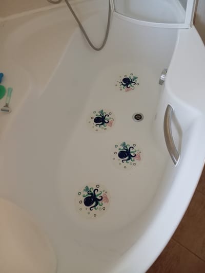 The image shows the interior of a white bathtub with four circular, octopus-themed anti-slip stickers adhered to the bottom. The stickers are light-colored with dark blue octopuses, pink coral, and green seaweed designs. A silver faucet and drain are visible in the center of the tub. A silver grab bar is mounted on the right side of the tub. On the left side of the tub, a razor and a green and blue bath accessory are present. The bathtub appears to be in a bathroom setting, with a glimpse of a brown tiled floor at the bottom right of the frame. The location is not specified in the image, and therefore, it cannot be confirmed to be in Baden, Austria.