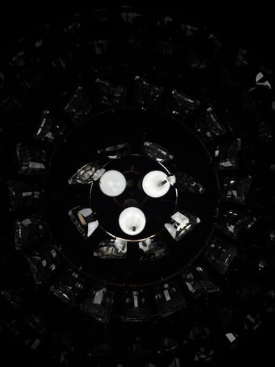 The image provides an upward-looking, overhead view of an illuminated chandelier, set against a stark, black background. In the center, three bright, round, white light bulbs glow, arranged in a circular cluster. These bulbs appear to be mounted on a dark, reflective base, possibly a mirror or polished surface, which is also circular.
Surrounding the central lights are numerous clear, faceted crystal or glass elements. These elements are arranged in concentric rings and are attached to a thin, circular metallic framework, likely made of brass or a similar golden-toned metal. The individual crystal pieces are somewhat rectangular or block-like, with cut surfaces that catch and refract the light from the bulbs, creating subtle reflections and glints.
The deep black background strongly suggests a dark environment, possibly an interior at night or a studio setup designed to isolate the chandelier. There are no people, visible activities, or discernible text within the image. The overall impression is one of elegance and sophisticated design, highlighting the chandelier's intricate details and luminous quality. While the context of Griboyedov, Armenia, is provided, there are no visual cues within the image to confirm this specific location.
