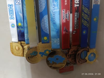 The image shows a collection of medals hanging on a wall. The medals have ribbons with various designs, including the flag of Kazakhstan and traditional Kazakh patterns. Some of the medals are gold, while others appear to be bronze or silver. Text on the ribbons and medals includes "QAZAQSTAN REPLIKASY", "CHAMPIONAT", "KICKBOXING ALMATY - 2025", "NAIZA 99 ROUND", and "SAQ PRO TEAM". There is also text in Cyrillic and what appears to be a date "27.06.2026 21:30" in the bottom right corner, indicating the image was taken on this date and time. The location is specified as Qaskeleñ, Kazakhstan. The medals are likely awards for athletic or other achievements. The background is a plain, light-colored wall.