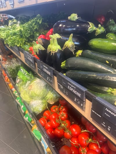The image shows a produce section in a store, likely a supermarket. The produce is arranged in bins with price tags indicating origin and price per kilogram. Visible produce includes tomatoes on the vine, zucchini, eggplant, and lettuce. Some of the produce has packaging. The price tags indicate that some of the produce originates from Spain and Belgium. The specific city and location are not discernible from the image.