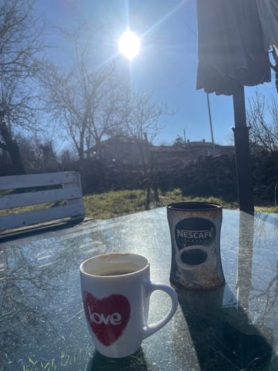 The scene depicts an outdoor setting in Topchii, Bulgaria, on a clear, sunny day. In the immediate foreground, a reflective glass tabletop occupies the lower portion of the frame. Resting on this table are two objects: a white ceramic mug with a red heart design enclosing the word "love," containing a dark liquid (likely coffee) with some residue adhering to its interior rim; and an empty, rusted Nescafé Classic instant coffee can, dark blue and silver branding partially obscured by corrosion. Strong sunlight from the upper right creates lens flare, reflections on the glass surface, and distinct shadows cast by the mug and can.
Beyond the tabletop, a low-lying, white wooden structure or bench is visible on the left. The midground features a patch of green grass, followed by a substantial mound of dark earth or debris. Overlooking this, several deciduous trees with bare branches are prominent, indicating a late autumn, winter, or early spring season. In the far right midground, the dark pole and folded fabric canopy of a garden umbrella are visible. In the background, partially obscured by the trees and terrain, are several residential buildings or houses with visible rooftops, suggesting a village environment. The sky is bright blue and clear, with the sun positioned high, producing a prominent starburst flare. No individuals are present in the image, and no active interactions are depicted.