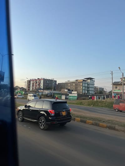 An outdoor, eye-level street scene in Biashara ward, Kenya, captured under a clear, light blue sky transitioning towards a lighter hue at the horizon. A black Subaru Forester SUV, bearing license plate KDJ 751A, occupies the foreground, traveling away from the viewer on a paved road with a yellow and black striped curb. In the mid-distance on the left, a white van and a white sedan are visible on the road. On the right side, further down, an orange three-wheeled auto-rickshaw is present.

The roadside to the right features a strip of unpaved ground, hosting several green rectangular structures, one labeled "Safaricom m-pesa." Adjacent to these, a linear arrangement of approximately ten mannequins, clothed in various garments, indicates an outdoor retail display. A group of about 8-10 individuals is gathered near these mannequins, facing them or conversing. Another solitary individual stands near the white van on the left.

In the background, multiple multi-story buildings populate the skyline. A prominent multi-story residential building with red-colored balconies is situated left of center. To its right, another building appears to be under construction or renovation, featuring exposed concrete and a green-framed section. Utility poles with numerous overhead wires are distributed across the scene. The left edge of the image is partially obscured by a vertical blue element.