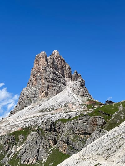 A towering, jagged rock formation dominates the center of the image, its weathered grey and tan surfaces stark against a vibrant, cloudless blue sky. The mountain's lower slopes are covered in scree and patches of green vegetation, with a winding path visible in the foreground. To the right, a cluster of buildings with dark roofs sits nestled on a grassy incline near the summit. The scene is set in the Dolomite mountains, likely near Colle Santa Lucia, Italy, a region known for its dramatic peaks and alpine scenery. The image captures a sense of rugged natural beauty and the isolation of a mountain refuge.