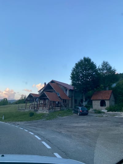 This is a photograph of a roadside establishment in Praga, Montenegro, captured during daylight hours. The image features a building with a red tile roof, possibly a restaurant or lodge, situated on a grassy slope adjacent to a paved road. In front of the building, there are covered outdoor seating areas with wooden tables and chairs. A dark-colored car is parked in the gravel area in front of the main building. To the right of the main structure, a smaller, unfinished concrete building with a red tile roof stands, appearing to be an outdoor oven or barbecue pit. Tall trees are visible behind the buildings, and the sky is a clear blue with some scattered clouds on the horizon. The road in the foreground has white dashed lines marking its lanes.
