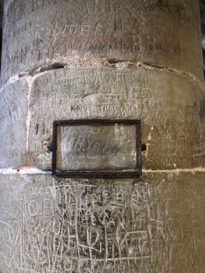 Close-up of a rough, light-colored stone column or wall, covered in carved graffiti. A dark-framed sign or display case is affixed to the stone, partially obscuring the carvings. The visible text inside the frame reads "BYRON". The photograph depicts graffiti attributed to Lord Byron in Château de Chillon, Veytaux, Switzerland. The stone surface shows signs of age and wear. Some carvings are shallow and faint, while others are more pronounced.