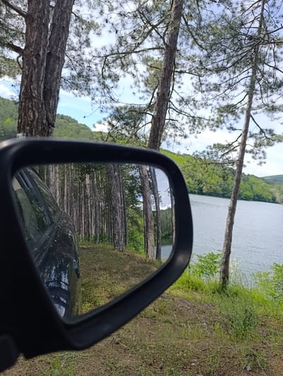 The image captures a serene lakeside view, reflecting a dense pine forest in the side-view mirror of a car. The mirror shows a reflection of tall, slender pine trees on a dirt path, with the dark exterior of the car visible in the foreground. Beyond the reflection, the actual scene unfolds with a calm, blue lake to the right, bordered by lush green hills covered in trees. The sky above is a mix of white clouds and blue. The car appears to be parked on a dirt road or trail next to the lake, surrounded by dry grass and pine needles. The location is identified as the dam of Georgi Dimitrov, near the village of Donyo Marinini, in the municipality of Nevestino, Bulgaria. There are no people or animals visible in the image.