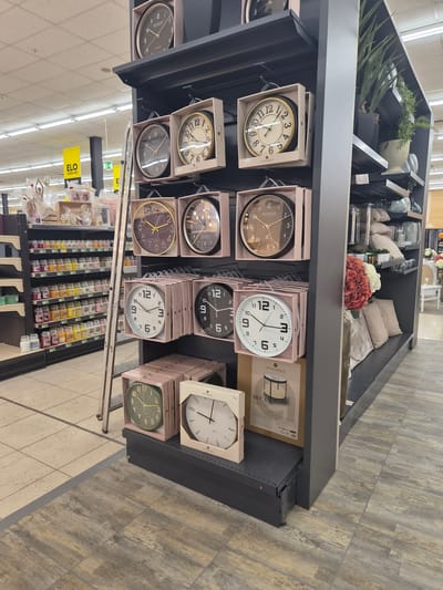 The image displays a retail setting, likely a home goods store, with a focus on clocks and decorative items. The scene is well-lit, suggesting an indoor environment during daytime hours. The primary subjects are various wall clocks presented in their packaging on shelves. These clocks feature different designs, including classic analog faces with Roman and Arabic numerals, as well as more modern, minimalist styles. Some clocks have ornate gold or silver-toned casings, while others are simpler in white or black.
Among the clocks, there are decorative items such as artificial flowers in shades of pink and red, and potted plants, suggesting these are part of a display for home decor. The shelves themselves are dark gray, contrasting with the pale pink packaging of the clocks. In the background, there are glimpses of other merchandise, including what appear to be candles or small jars of scented products, arranged on lower shelves. The flooring is a mix of light-colored tiles and wood-look planks.
Visible text includes brand names like "atmosphera" and "WESTMINSTER", along with product designations such as "MECA". A prominent yellow sign with the text "ELO ODER NIE!" is also visible, which appears to be a promotional or directional sign within the store. The overall impression is a tidy and organized retail display, aimed at attracting customers interested in home furnishings and accessories. There are no people visible in the image.