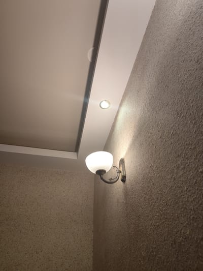 This media file depicts an interior scene, likely a hallway or room, with a textured beige wall and a stepped ceiling. A wall-mounted light fixture with a frosted glass shade and an ornate metal base illuminates the wall. Above and to the left, a recessed ceiling light is also visible, casting a soft glow. The textured nature of the wall suggests a decorative finish, possibly wallpaper or a stucco effect. The lighting indicates it is likely evening or nighttime, as the interior lights are the primary source of illumination. No people or text are present in the image. The setting appears to be a residential or commercial interior space in Sillamäe linn, Estonia, characterized by its muted color palette and functional yet decorative lighting. The scene conveys a sense of quietude and simple elegance.