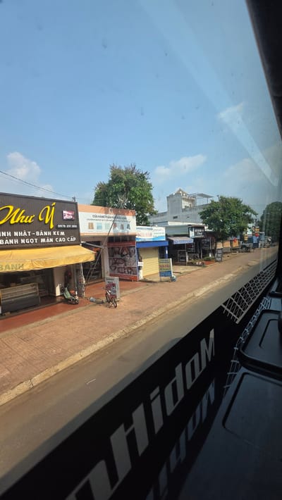 Captured from inside a vehicle, the image portrays a street scene in Xuân Long, Vietnam. The camera is positioned high, offering a view out the window and a portion of the vehicle's interior. Outside, a clear blue sky speckled with wispy clouds meets the rooftops of closely packed buildings lining the street. The businesses display signs in Vietnamese, advertising a variety of goods and services, including baked goods, home furnishings, and hair salons. Bicycles and scooters are parked on the sidewalk in front of the shops. The vehicle's interior, visible in the foreground, includes part of the dashboard. The lighting is bright, suggesting a sunny day.