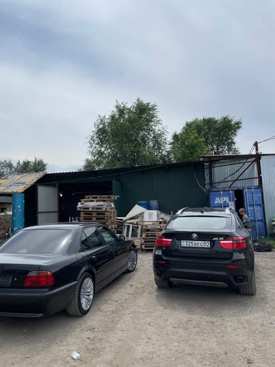 Two black BMWs are parked in an outdoor industrial or storage area. The car on the left appears to be a sedan, and the car on the right is an SUV. In the background, there is a long, dark green building with an open section revealing a darker interior. To the left of the building, there are blue barrels and a damaged roof. To the right of the building, a large blue shipping container is partially visible, with a person standing near its open door. Piles of wooden pallets are stacked near the building. The ground is a mixture of dirt and gravel. Trees are visible behind the building, and the sky is overcast with some light clouds. The scene is located in the village of Kenen Azirbayev, Kazakhstan.