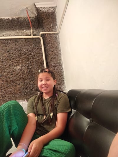A young girl with braided hair, adorned with star-shaped clips, sits on a black leather couch in what appears to be an indoor setting. She is wearing a dark green t-shirt and vibrant green pleated pants. Her expression is cheerful as she looks towards the camera. The background features a wall covered in small, dark, rounded stones, with exposed pipes running along the top. The lighting suggests it is daytime, though the indoor environment makes it difficult to ascertain the exact time of day or weather conditions. The girl is holding a phone or similar device in her lap. The overall scene conveys a casual and relaxed atmosphere.