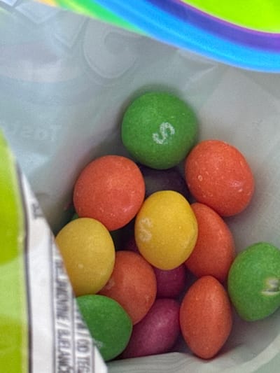 The scene depicts a close-up, high-angle view looking into an open, flexible bag containing numerous multi-colored, spherical to oval-shaped candies. The candies are primarily green, orange, yellow, and purple/magenta. A distinct white "S" is visible on the surface of one green candy located centrally in the upper portion of the image. Other candies show minor surface imperfections and white speckling. The interior of the bag is light gray or white. A blurred, rainbow-colored section (blue, purple, green, yellow) is visible along the top edge, likely part of the bag's opening or design. On the left side, a partially torn edge of the bag reveals a green outer layer and a white strip with fragmented text, including segments resembling "PEČIVINE" or "BLANCHE," indicating product labeling. The immediate environment is contained within the confines of the candy bag. This scene is located in the city of Cerovo, Montenegro.