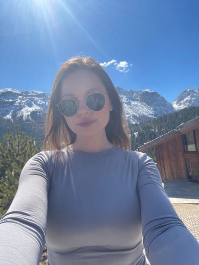 A selfie shows a woman wearing sunglasses, a long-sleeved gray shirt, and standing outside with mountains in the background. Snow is visible on the mountain tops. Some trees and a wooden building are also visible. The photograph was taken in the city of Sappada / Plodn / Sapade, Italy.