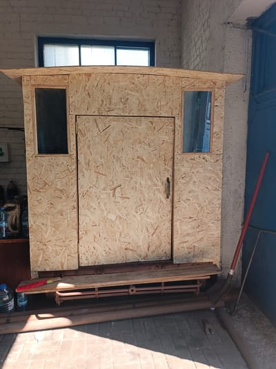 This media file shows a partially constructed wooden structure, possibly a shed or a small shelter, made from oriented strand board (OSB). The structure has a simple, rustic appearance. There is a door in the center, with a metal handle. Two small windows, one on each side of the door, are visible. The top of the structure has an overhanging roof, also made of OSB. The structure appears to be situated in an outdoor or semi-outdoor setting, possibly a workshop or yard, with a brick wall in the background and concrete floor with some metal pipes and a shovel lying nearby.
The sunlight suggests it is daytime. The overall impression is one of construction or renovation in progress. There is no visible text in the image. The location context provided is Vuhlehirsk, Ukraine.