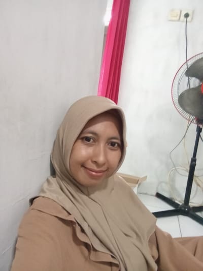 An indoor photograph captures a single adult female, appearing to be of Southeast Asian descent, positioned centrally and looking directly at the camera with a slight smile. She wears a light brown hijab and a light brown long-sleeved garment, appearing to lean against an off-white or light grey wall. To the left in the background, a vertical strip of pink fabric is visible. To the right, a black standing electric fan with a red-rimmed circular grille and dark blades is present. Above the fan, two white electrical outlets are mounted on the wall. A light-colored cardboard box is partially visible on the floor behind the subject. The floor surface appears light-colored, possibly tiled. The specific location in Bulukandang, Indonesia, cannot be confirmed from the visual information in the image.