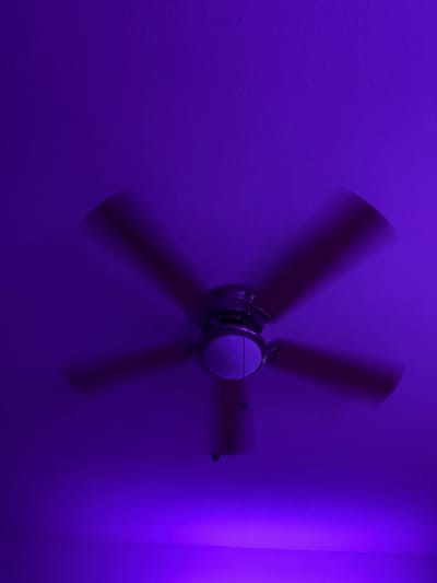 An overhead, low-angle shot shows a ceiling fan in motion, illuminated by a vibrant purple light. The fan has a silver motor housing and five dark-colored blades that are blurred due to the long exposure. Two pull chains hang from the fan's motor. The ceiling is painted a solid, textured purple, with the light casting a brighter, more saturated purple glow on the lower portion of the image, suggesting the light source is positioned below the fan and angled upwards. The scene is static and devoid of people.