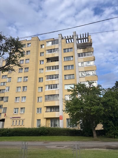 A medium shot captures the side of a tall, yellow apartment building under a cloudy sky. The building features numerous windows, some with balconies. To the left of the building, a large tree with green leaves partially obstructs the view. In the foreground, a low green hedge runs along the base of the building, and beyond that, a paved sidewalk and a grassy area are visible. A chain-link fence separates the grassy area from a road or pathway. The building has a sign that reads "НОТАРИУСЫ" in purple letters, along with what appears to be a logo. Another sign on the building shows a red square with the number "1" inside and text above it. Power lines are strung across the top of the image. The overall scene depicts a residential or commercial building in an urban or suburban setting.