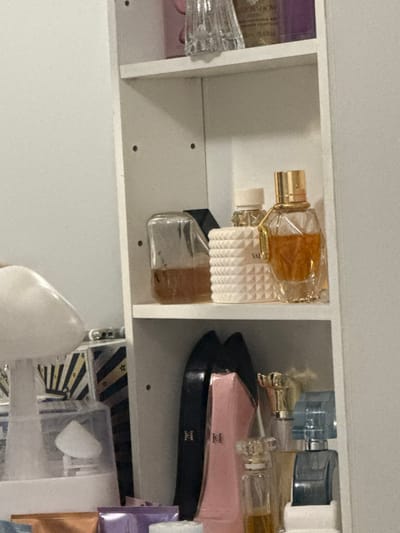 A white cabinet with multiple compartments holds various items, primarily perfume bottles. There are glass bottles with gold accents, a white studded bottle with a black base, and a pink bottle with a black label. Other items include a blue bottle, a clear plastic container with white contents, and decorative boxes with stripes and gold stars. The cabinet appears to be part of a vanity or storage area.