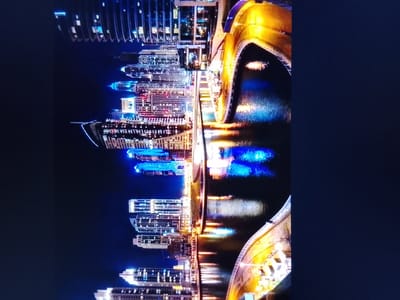 This is a vertically oriented, low-light, digitally captured image of a modern city skyline at night, reflected in water. The image is grainy and appears to be taken from a slight elevation, looking down at the cityscape. The buildings are illuminated with various colored lights, primarily white, blue, and yellow, creating a vibrant and dynamic scene. A prominent structure in the foreground is a curved, illuminated bridge or walkway with a golden hue, arching over the water. The water reflects the lights of the buildings and the bridge, creating a shimmering effect. There are no visible people or vehicles in the image. The location is identified as Safonovo, Russia, however, the architectural style does not suggest a typical Russian cityscape, and the visual elements are more indicative of a contemporary, affluent urban development, such as a harbor or waterfront district in a global city.