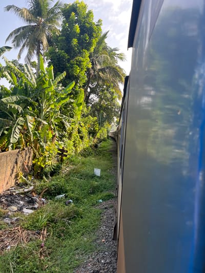 The image captures a view from a moving train alongside lush vegetation and a section of track. On the left, a concrete wall is visible, bordered by a pile of debris and overgrown green plants, including banana trees and palm trees. The train's metallic blue side occupies the right portion of the frame, reflecting the sky and surrounding trees. The train is on a track laid with dark gray ballast. A white marker with text is partially visible in the lower right quadrant. The environment suggests a rural or semi-rural setting in Maggona, Sri Lanka, with dense foliage typical of the region. No people are present in the image.